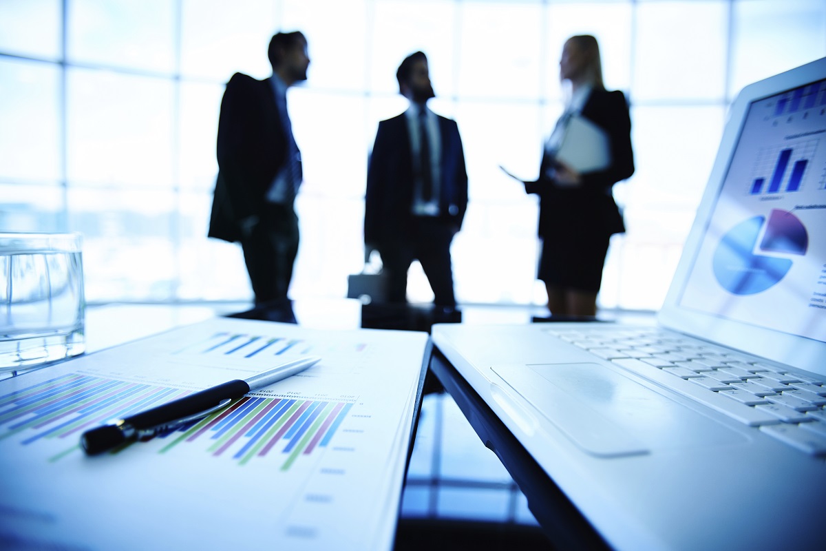 Laptop and financial document with pen at workplace on background of three business partners interacting